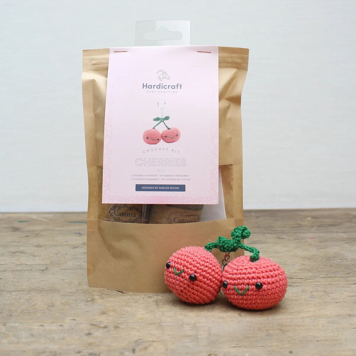 Cherries decoration crochet kit kraft bag packaging with pink labelling and completed crocheted cherries in front on wooden surface