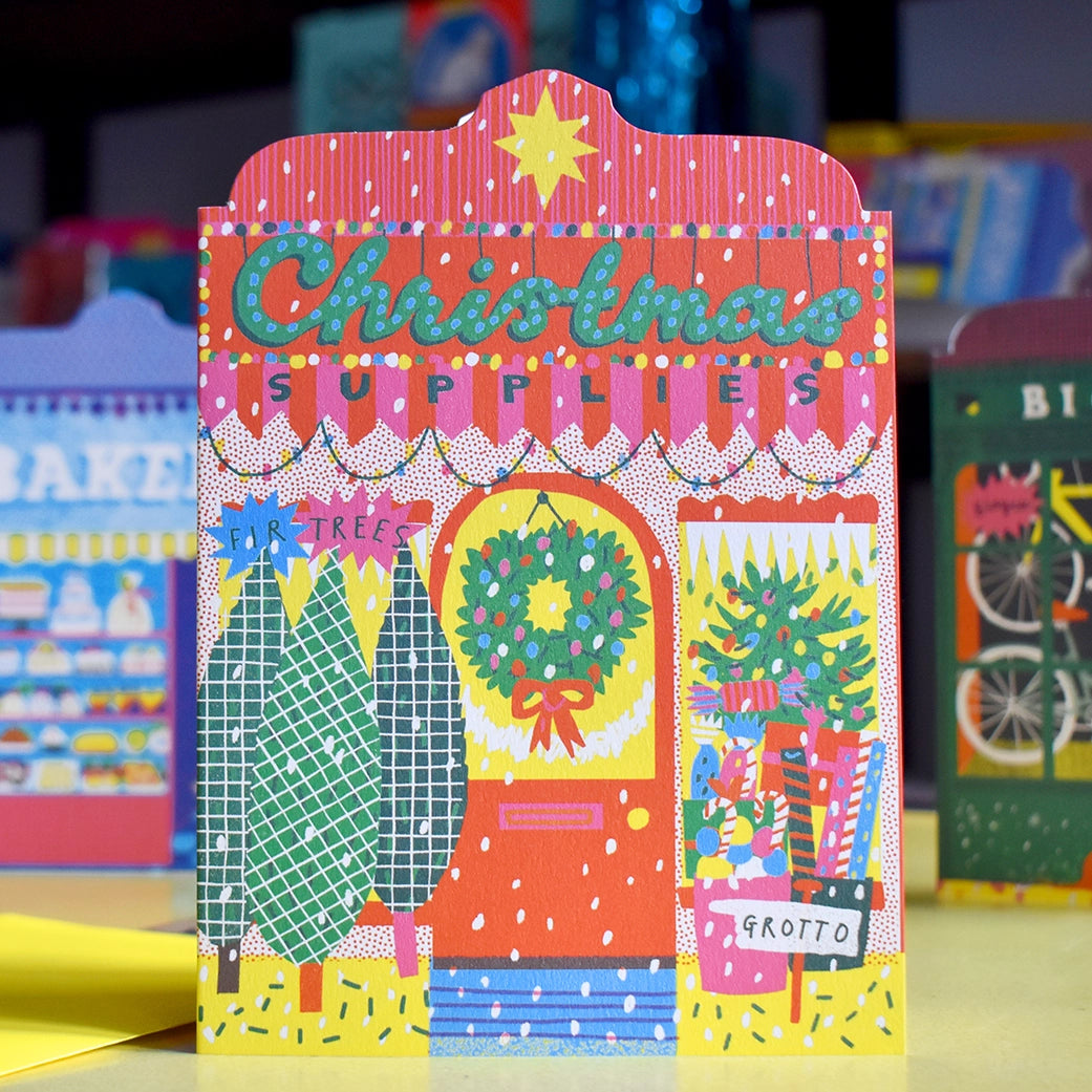 Illustrated note card featuring Christmas Supplies shop front decorated with artistic Christmas trees, wreath and wrapping paper in bright colours, set against a background of other artistic storefront cards