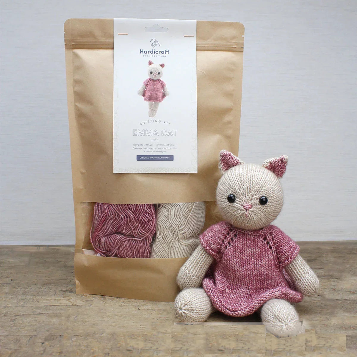 Knitted cat toy in a pink dress and kraft packaging with a window revealing pink and cream coloured yarn, on a wooden surface