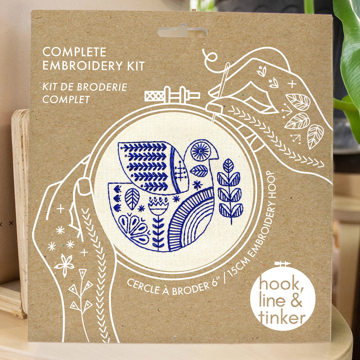Kraft packaging for a folk bird embroidery kit, showing illustrated hands holding a needle and thread around the design