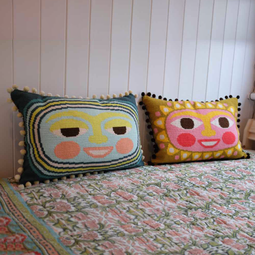 Two colourful needlepoint pillows with lunar and solar face designs on a floral-patterned bedspread