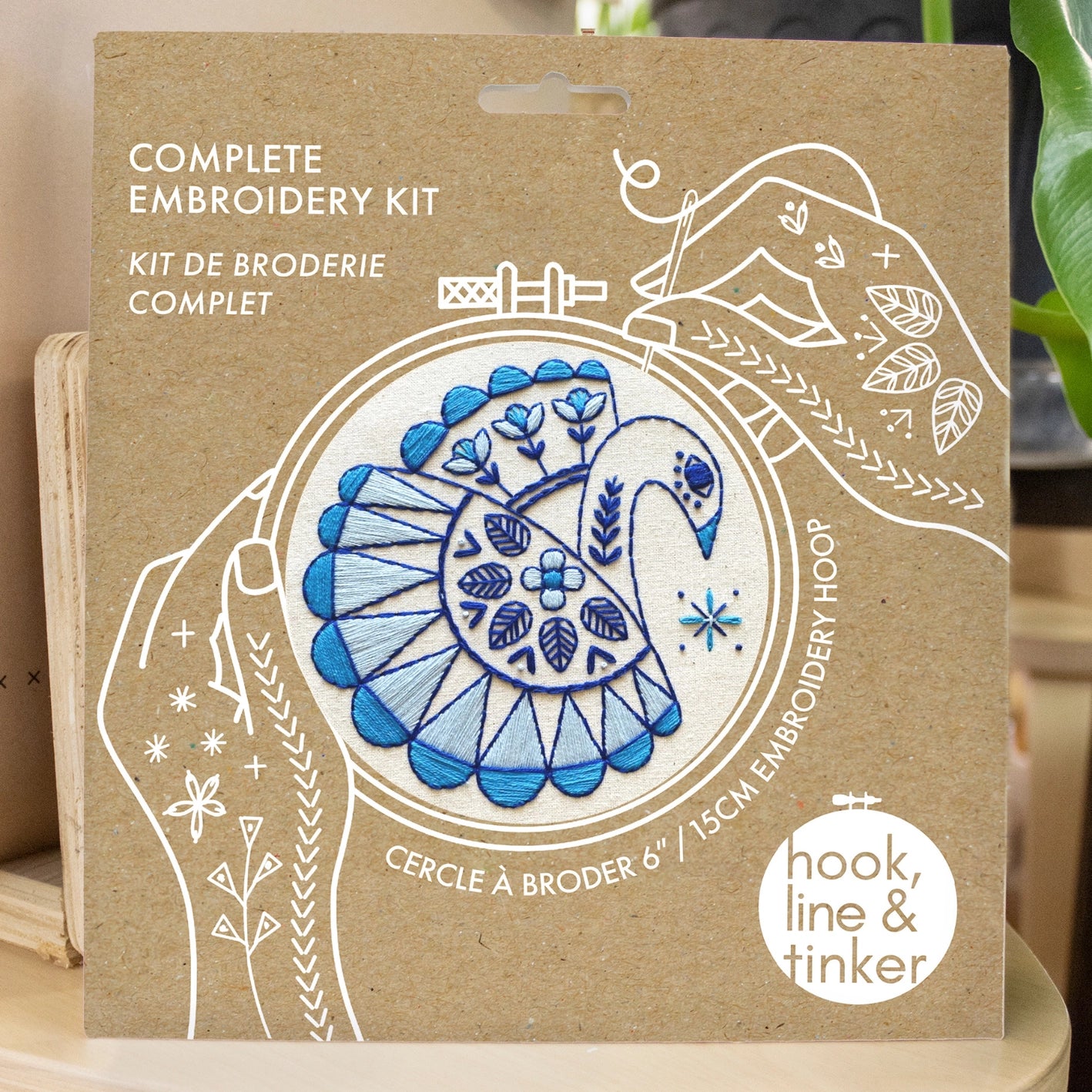 Kraft packaging for a swan embroidery kit, showing illustrated hands holding a needle and thread around the design