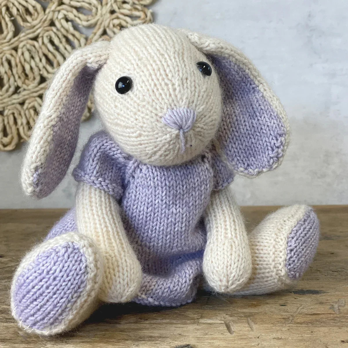 Knitted rabbit soft toy wearing a light purple dress sitting on a wooden surface