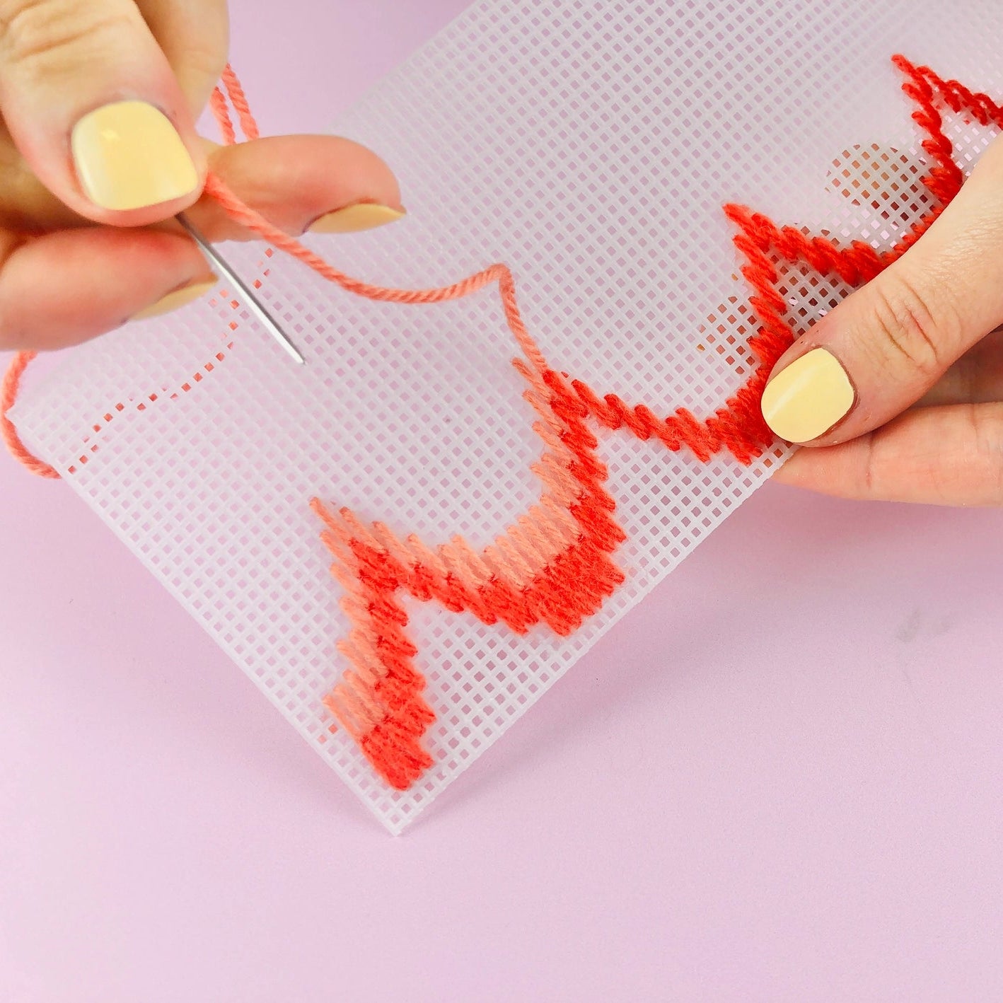 Close-up demonstrating stitching of bargello plant pot needlepoint, featuring orange and pink yarn and needle, and plastic needlepoint canvas