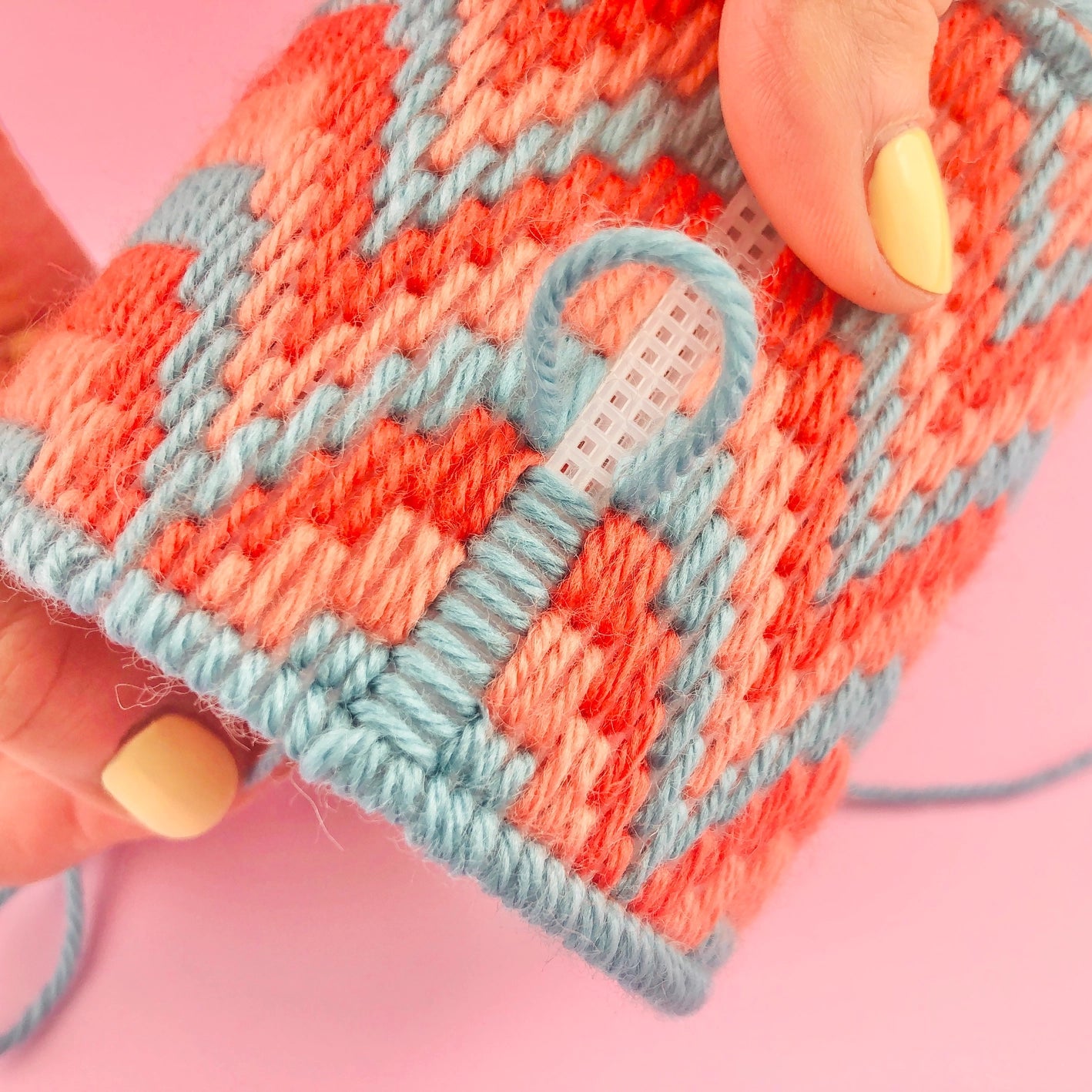 Close-up demonstrating stitching of bargello plant pot needlepoint, featuring orange and pink yarn and needle, and needlepoint canvas, demonstrating how to finish the pot