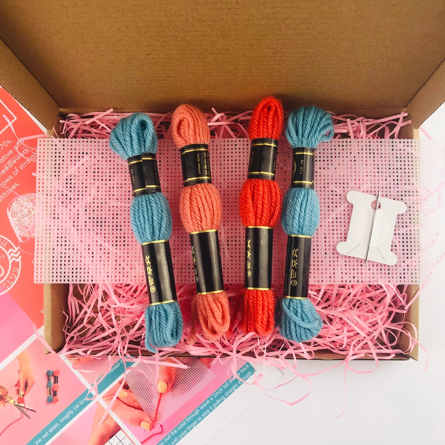 Close-up showing contents of bargello plant pot needlepoint kit, including orange and blue yarn, needle and plastic needlepoint canvas on a pink paper straw background
