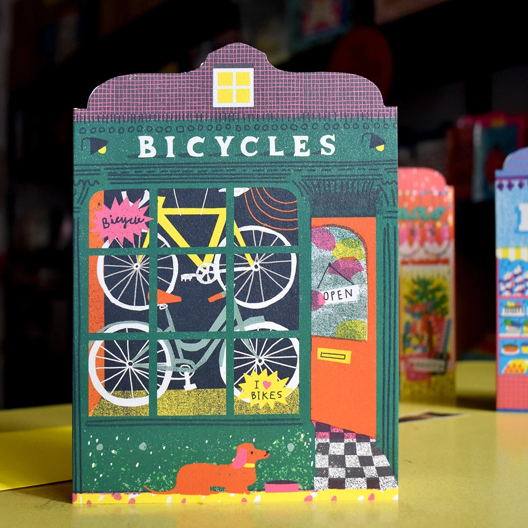 Illustrated note card featuring Bicycles shop front with bicycles hanging in the window, an open door and a dog in front, set against a background of other artistic storefront cards