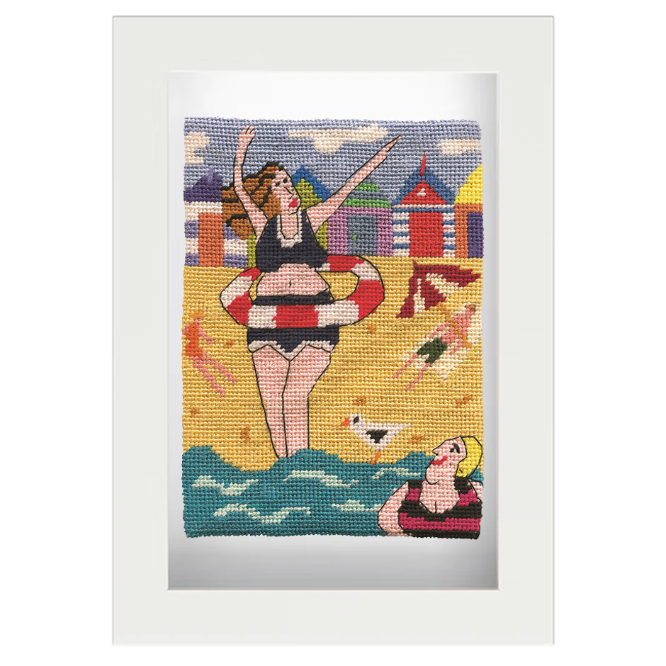 Framed needlepoint of women wearing swimsuits on a beach scene, with cabin huts in background