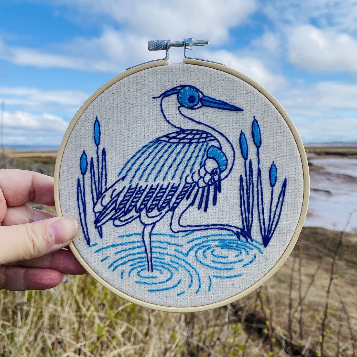 Finished blue heron embroidery held by hand against a coastal background