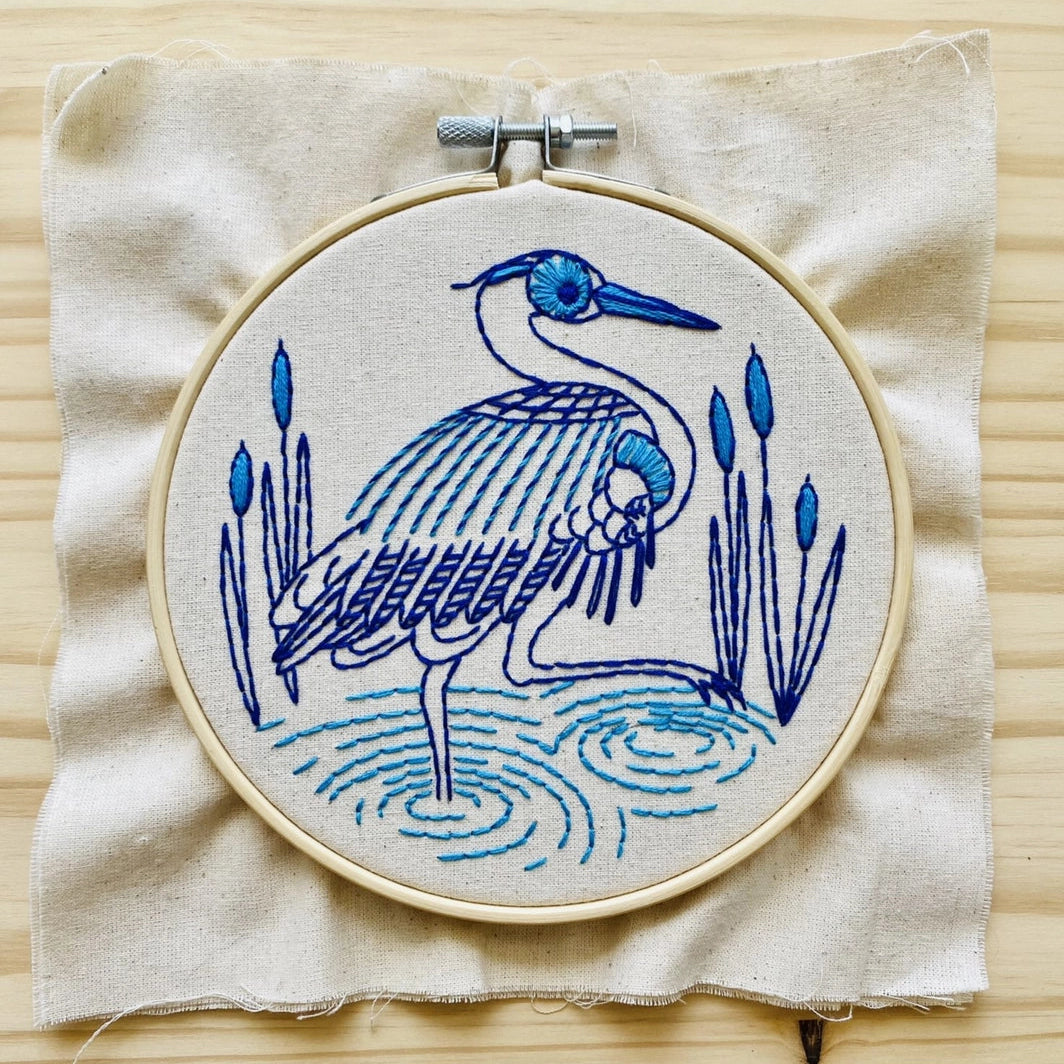 Finished embroidery of a blue heron on a square of unbleached calico fabric in a hoop on a wood background