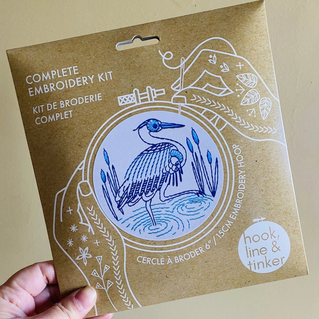 Hand holding kraft packaging for a blue heron embroidery kit, showing illustrated hands holding a needle and thread around the design