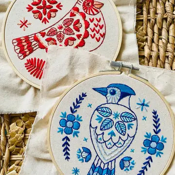 Embroidered design of a blue jay bird with floral patterns on unbleached cotton fabric, hoop-mounted and in-progress, against a background of a red cardinal bird embroidery