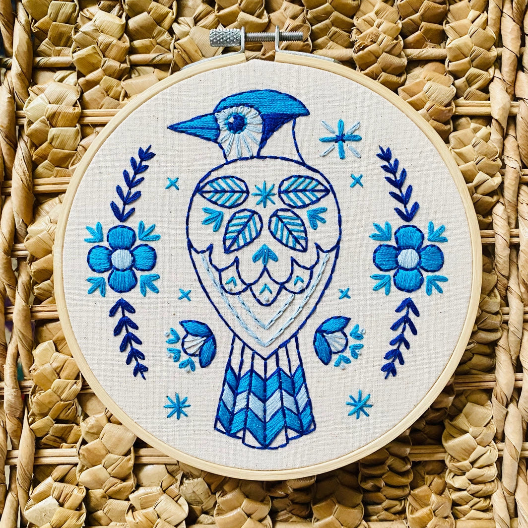 Completed embroidery of a blue jay bird with floral patterns on unbleached cotton fabric, hoop-mounted, on a straw background
