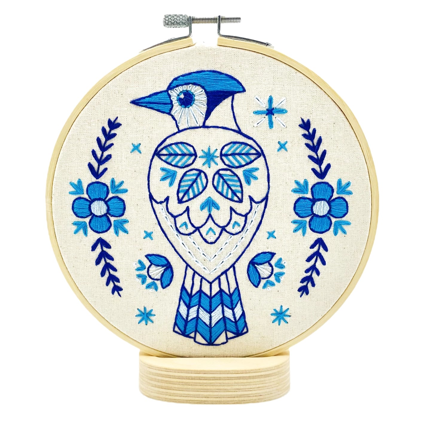 Completed embroidery of a blue jay bird with floral patterns on unbleached cotton fabric, hoop-mounted on a circular wooden base