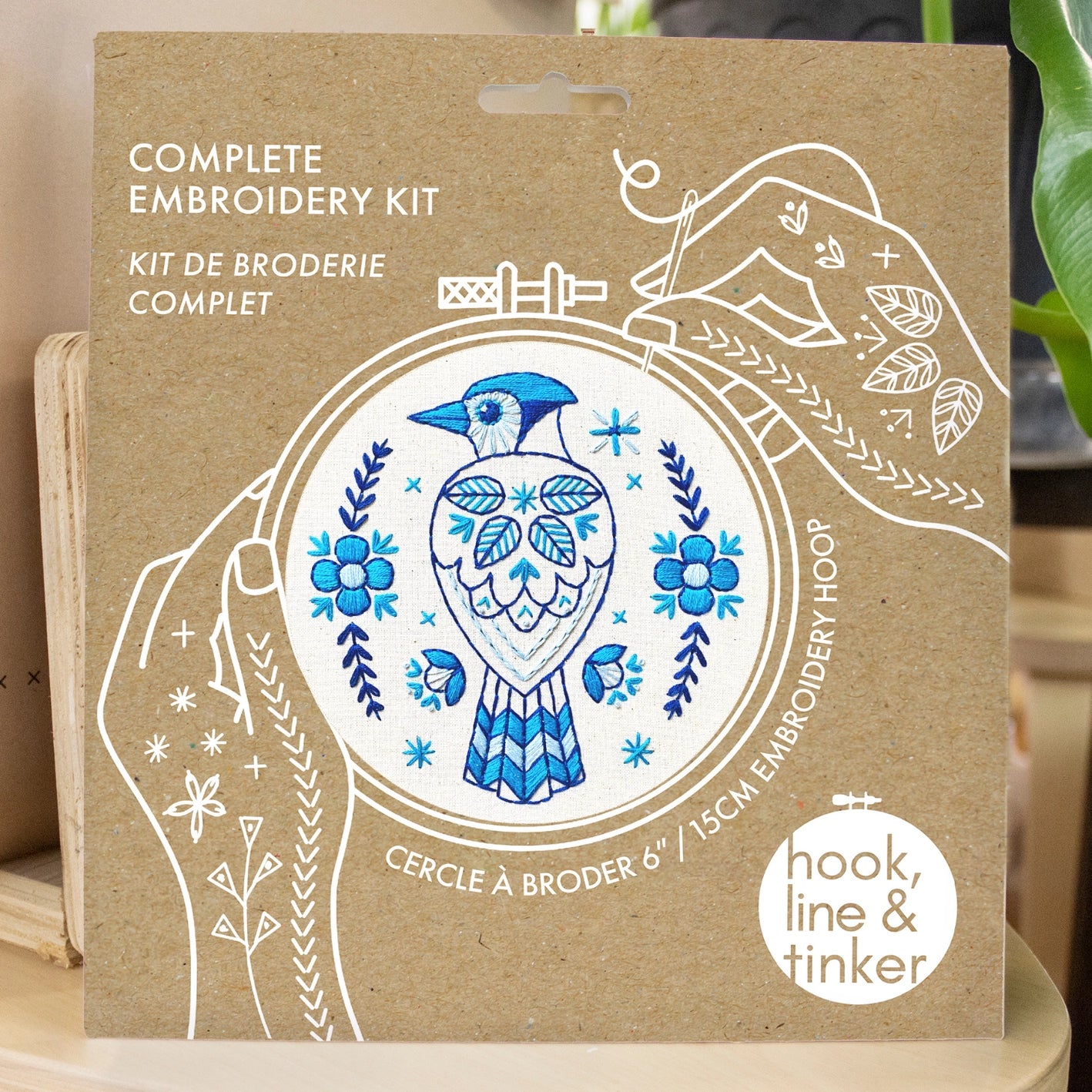 Kraft packaging for a blue jay embroidery kit, showing illustrated hands holding a needle and thread around the design
