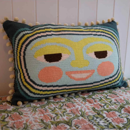 Decorative needlepoint pillow with a lunar face design in shades of blue, yellow, and orange on a floral-patterned bedspread