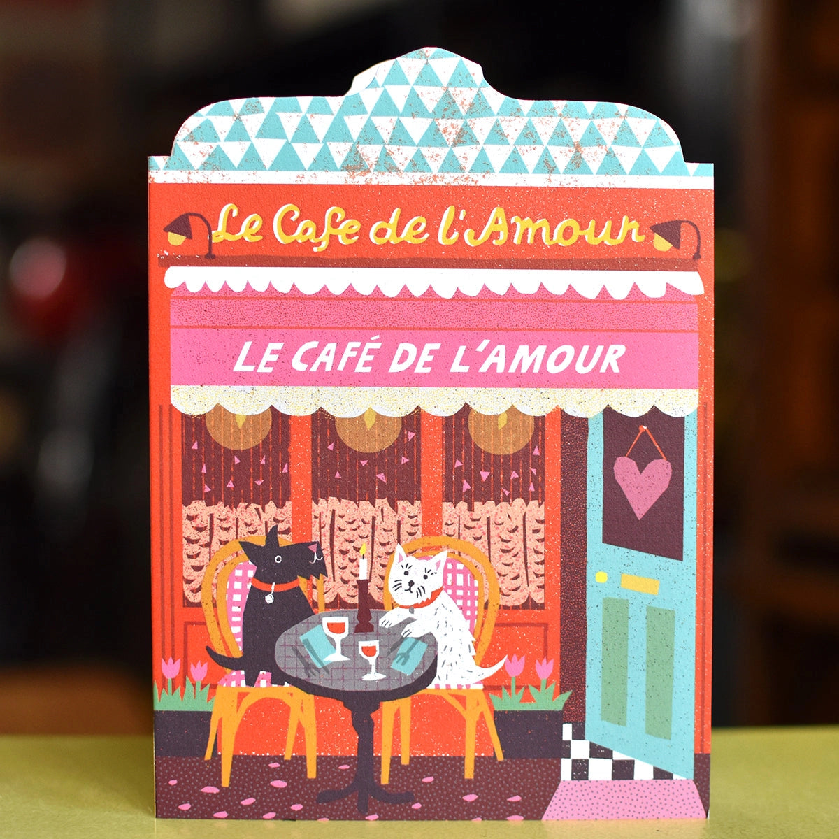 Illustrated card featuring Café de l'Amour, with café curtains, blue-green open door and heart sign, and two dogs  sitting at a table outside, set against a yellow and brown blurred background