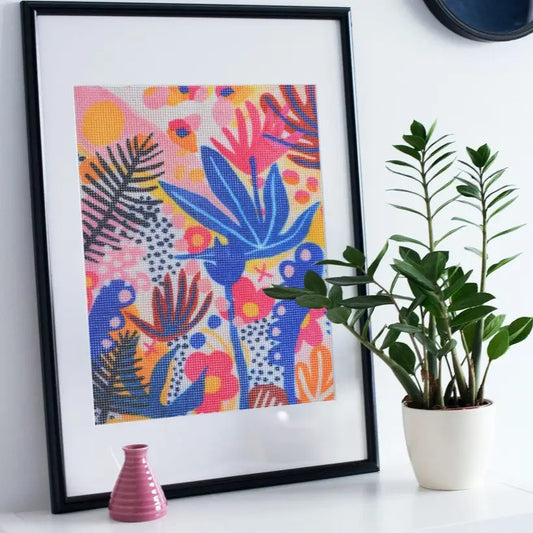 Framed needlepoint with a vibrant tropical floral design, displayed on a shelf with a potted plant and a small pink vase