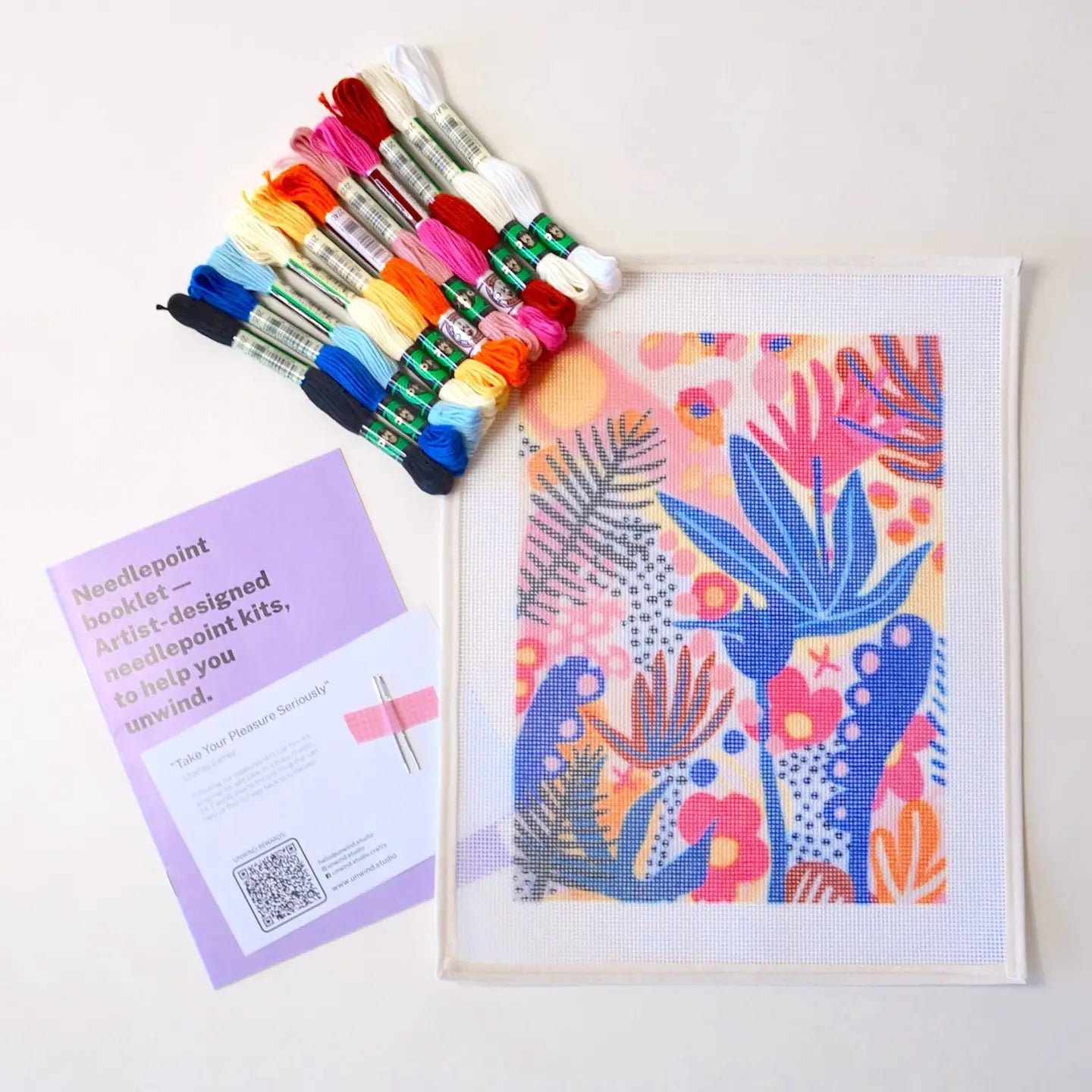 Contents of a needlepoint kit with a vibrant tropical botancial design, including a printed canvas, coloured threads, two tapestry needles, and instruction booklet