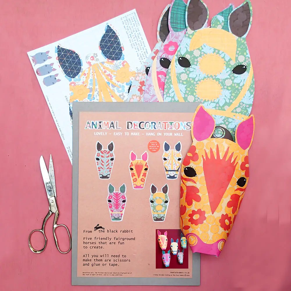 Contents of the carousel horse paper kit, including five fairground horse cards in different colours, an instruction sheet and a pair of scissors on pink background