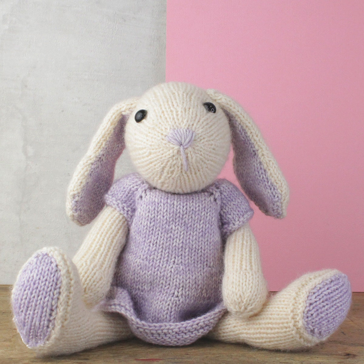 Knitted bunny toy wearing a purple dress on a wooden surface with a pink and white background