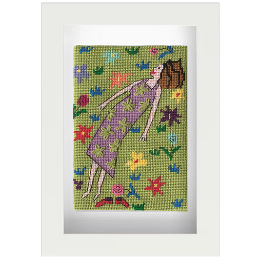 Needlepoint postcard of a woman lying in a colourful field watching clouds, on a green background, framed in white