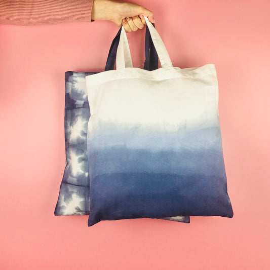 Two tote bags with a blue and white pattern, one featuring a blue to white gradient and the other with a blue and white tie dye pattern, held against a pink background