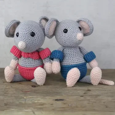 Two crocheted mice toys, one in a pink bikini and armbands and the other in a blue swimsuit and armbands, sitting on a wooden surface with a grey background