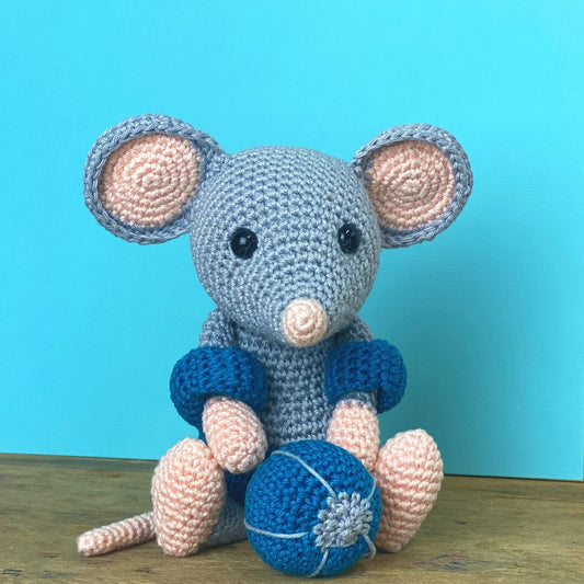 Crocheted mouse toy wearing a blue swimsuit and armbands, with a blue ball on a wooden surface against a blue background