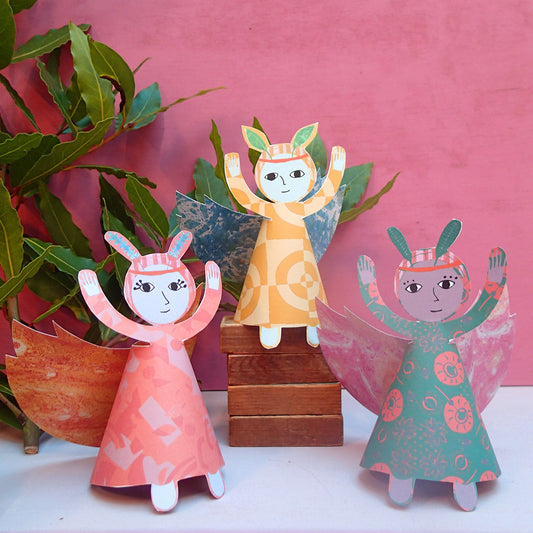 Three paper fairy angels in peach, green and yellow with contrasting wings, on a pink and white background with green foliage