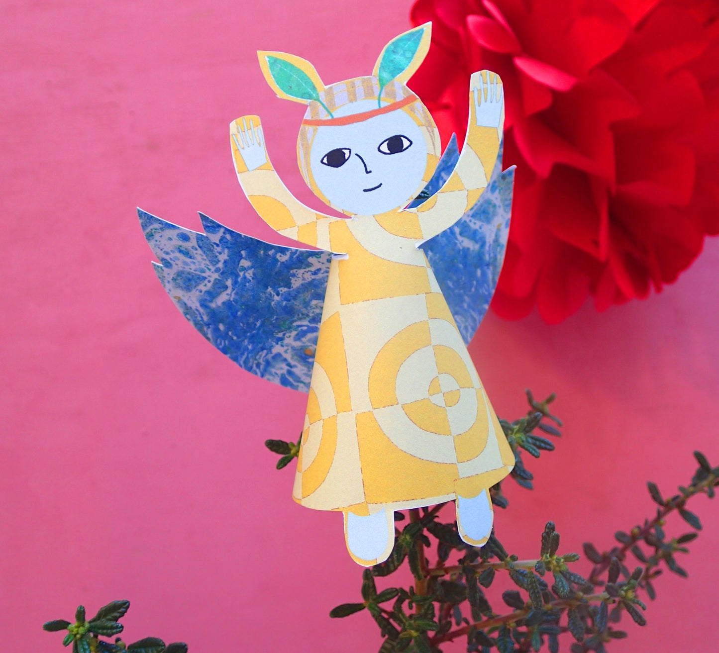 Close up of finished yellow festive fairy decoration with blue wings and green leaf tiara on pink background 