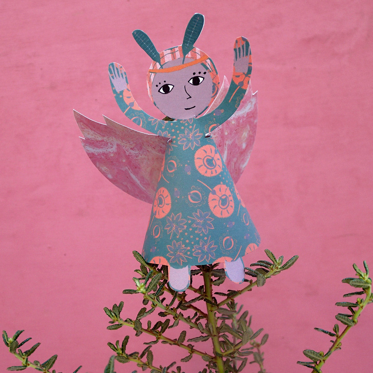 Close-up of finished green and orange festive fairy decoration with pink wings and green leaf tiara on pink background 