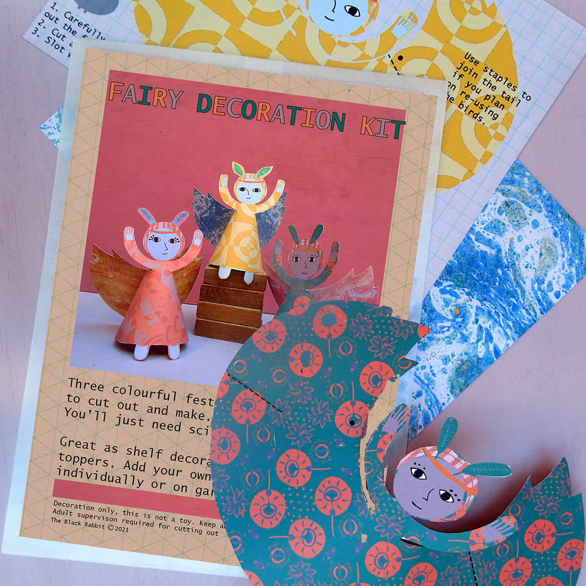 Close-up of fairy decoration kit kraft packaging with uncut cards in the background and a cut-out green and orange fairy in the foreground