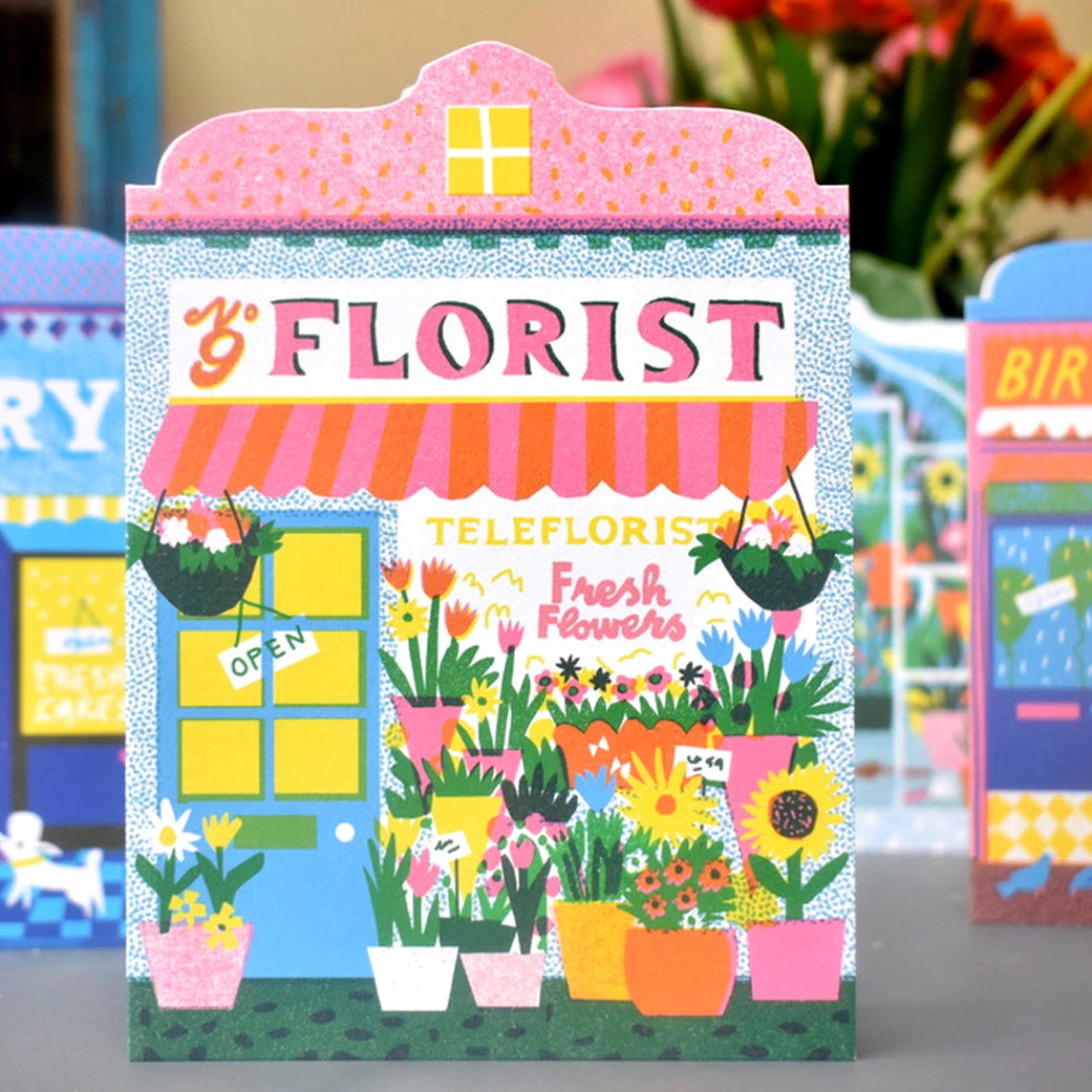 Illustrated note card featuring a colourful florist shop front with pots and vases full of flowers, with other cards in background