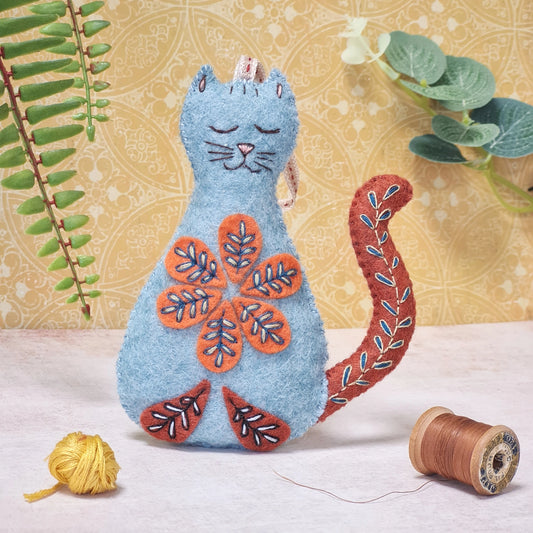 Completed folk-embroidered felt cat kit, with a blue body, orange details, and floral stitching, shown with crafting materials