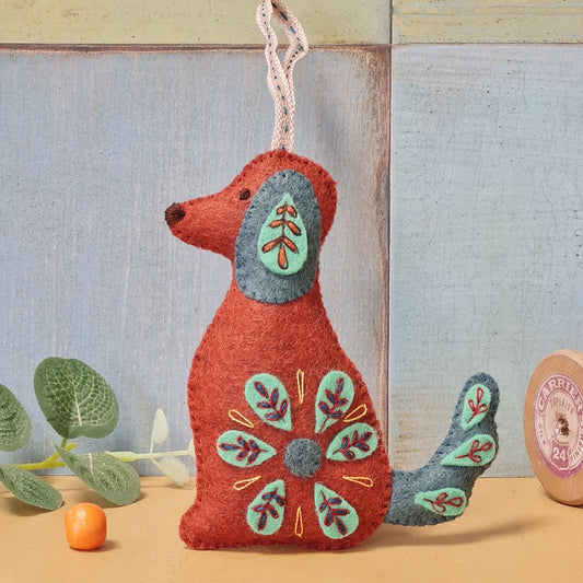 Completed folk-embroidered felt dog kit, with an orange-brown body, green and grey details, and floral stitching on a yellow and blue background