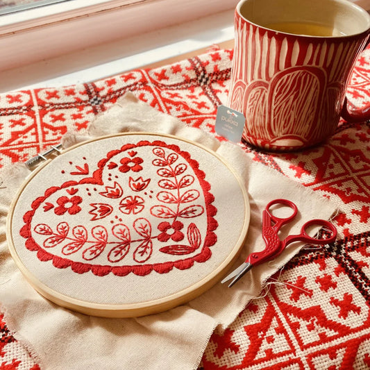 Embroidery hoop with red floral design on a patterned tablecloth with a mug and scissors