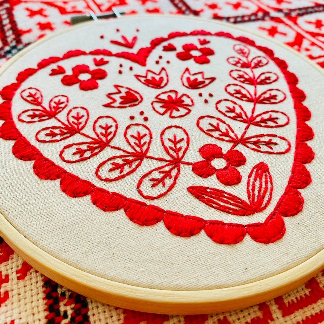 Folk-inspired embroidered red heart with floral patterns on unbleached calico, displayed on a red and natural needlepoint background