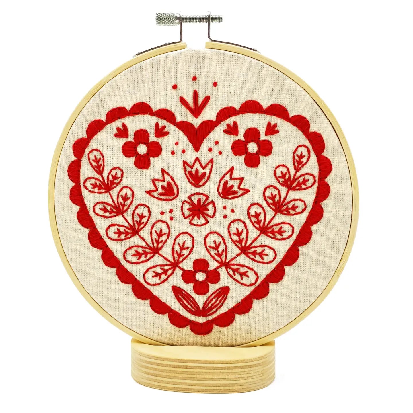 Folk-inspired embroidered red heart with floral patterns on unbleached calico, displayed in a wooden hoop on a small round stand
