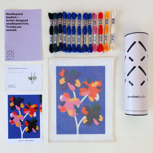 Contents of a needlepoint kit with a vibrant Four Flowers design, including a printed canvas, coloured threads, a photo of the original artwork, two tapestry needles, instruction booklet, and packaging tube