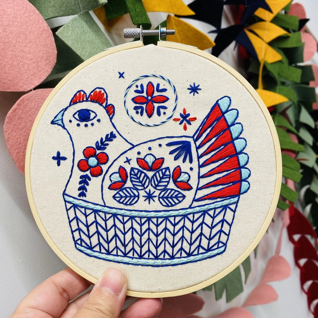 Hand holding a completed French hen embroidery in a colourful Scandi folk style, set against a bright background