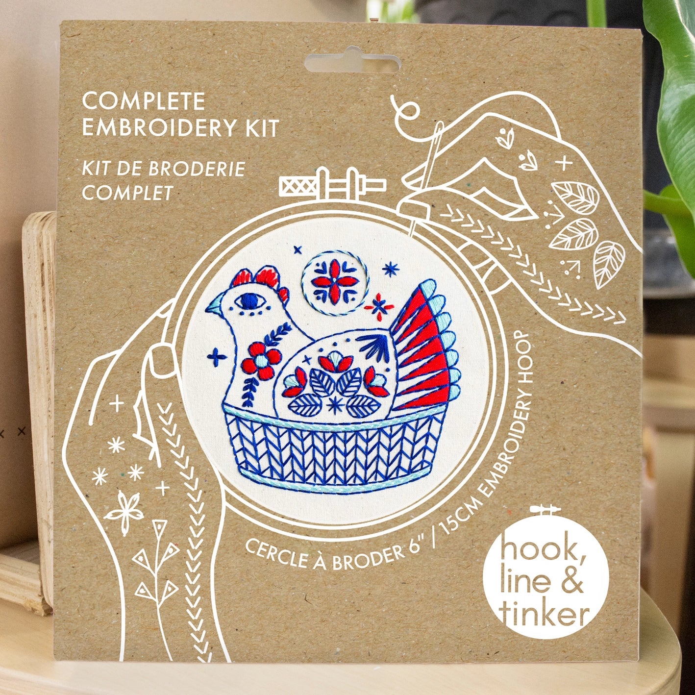 Kraft packaging for a French hen embroidery kit, showing illustrated hands holding a needle and thread around the design