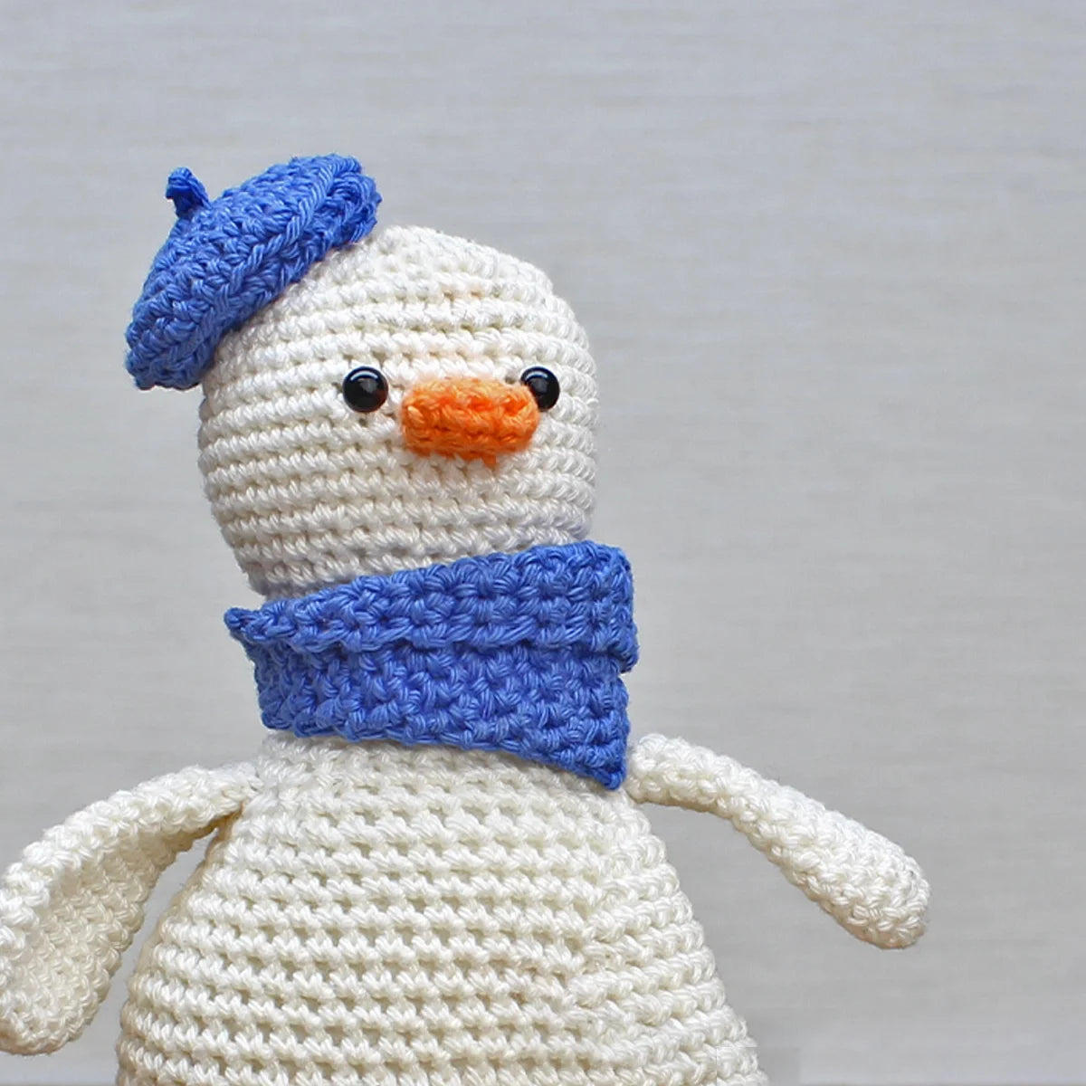 Close-up of crocheted goose toy with a blue hat and scarf on a plain background
