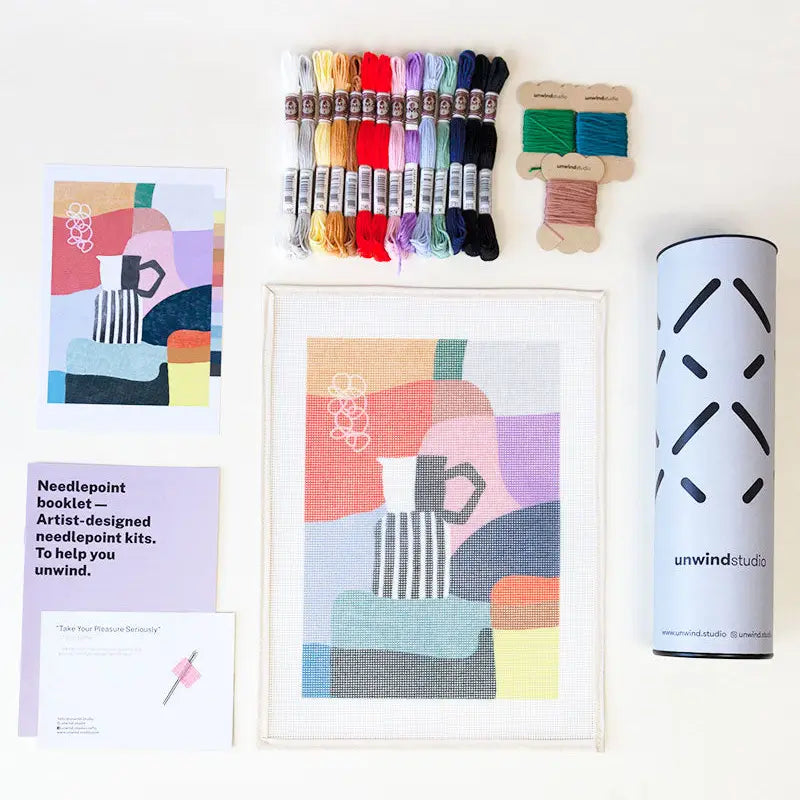 Contents of a needlepoint kit with an abstract coffee-themed design, including a printed canvas, coloured threads, a photo of the original artwork, two tapestry needles, instruction booklet, and packaging tube