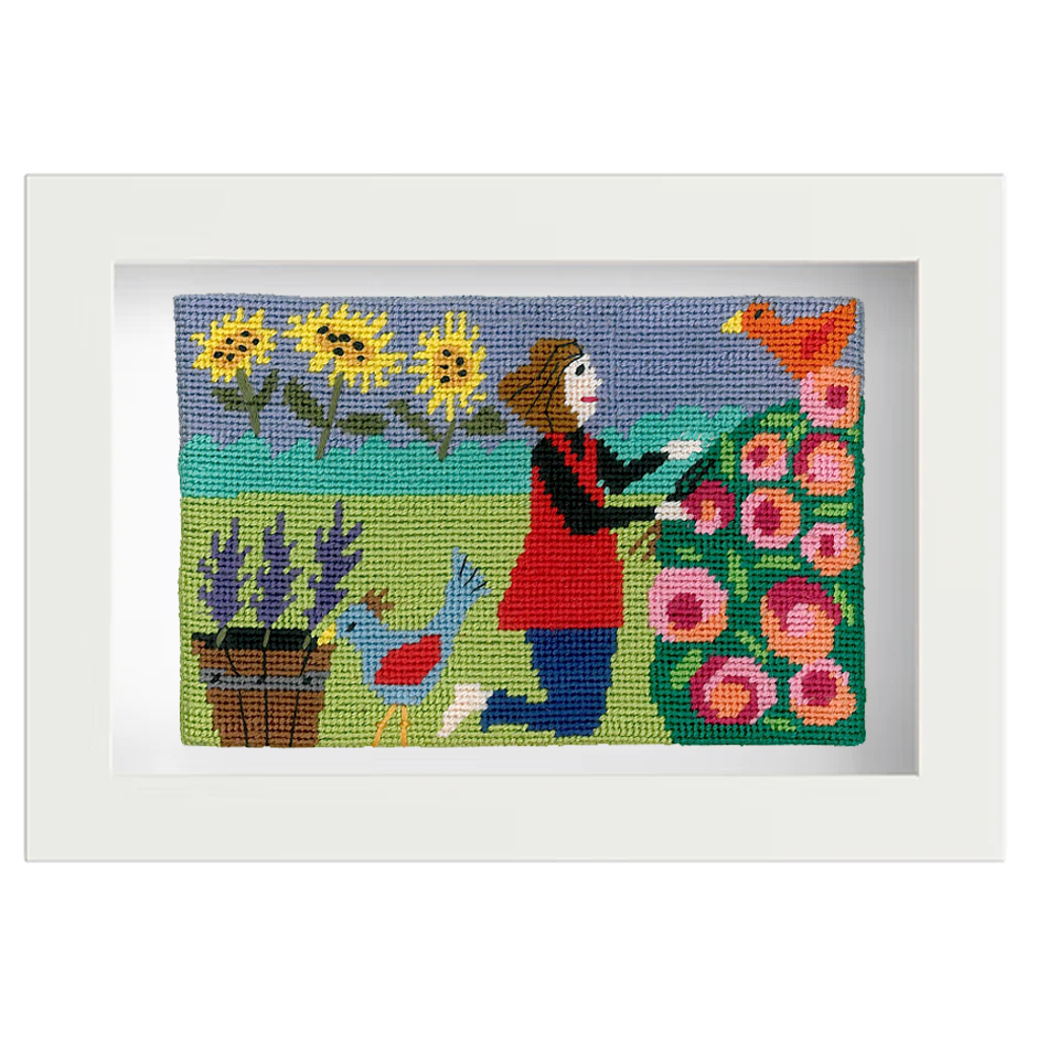 Framed, colourful needlepoint picture of a woman gardening with flowers and birds