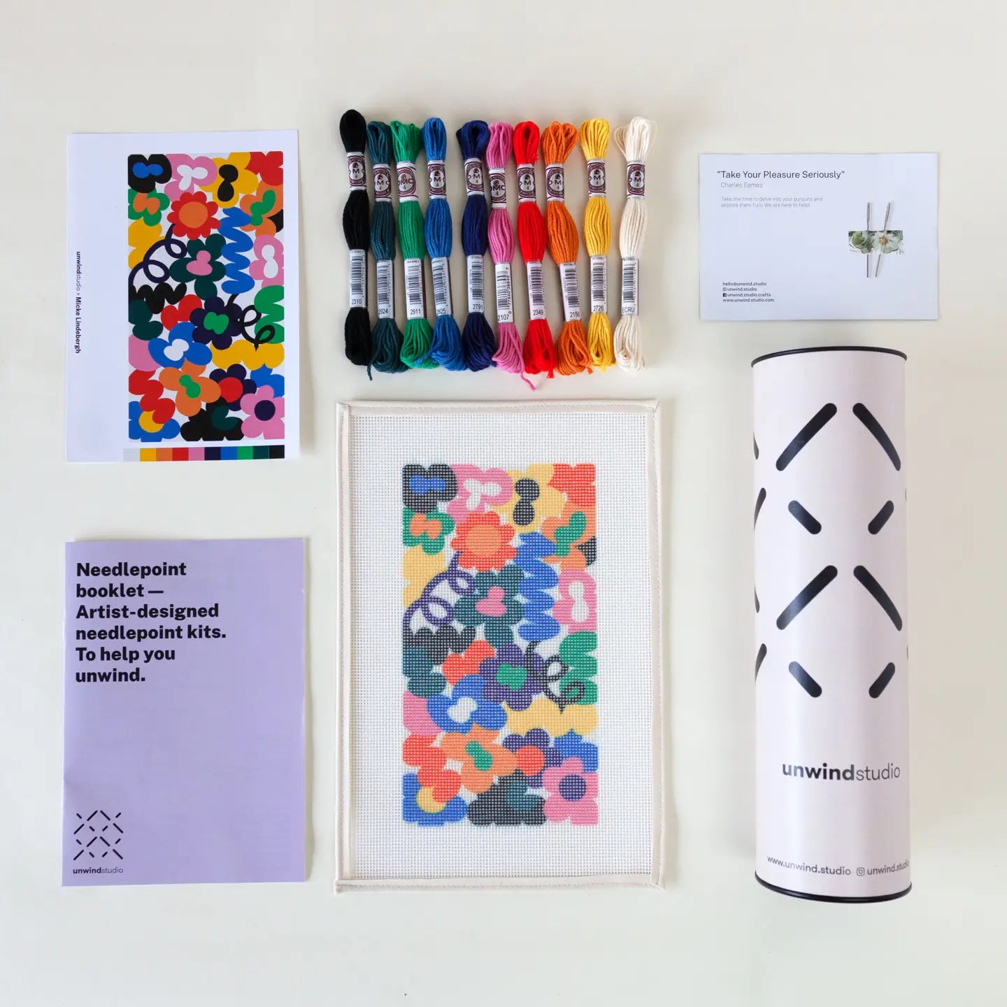 Contents of a needlepoint kit with a vibrant floral design, including a printed canvas, coloured threads, a photo of the original artwork, two tapestry needles, instruction booklet, and packaging tube