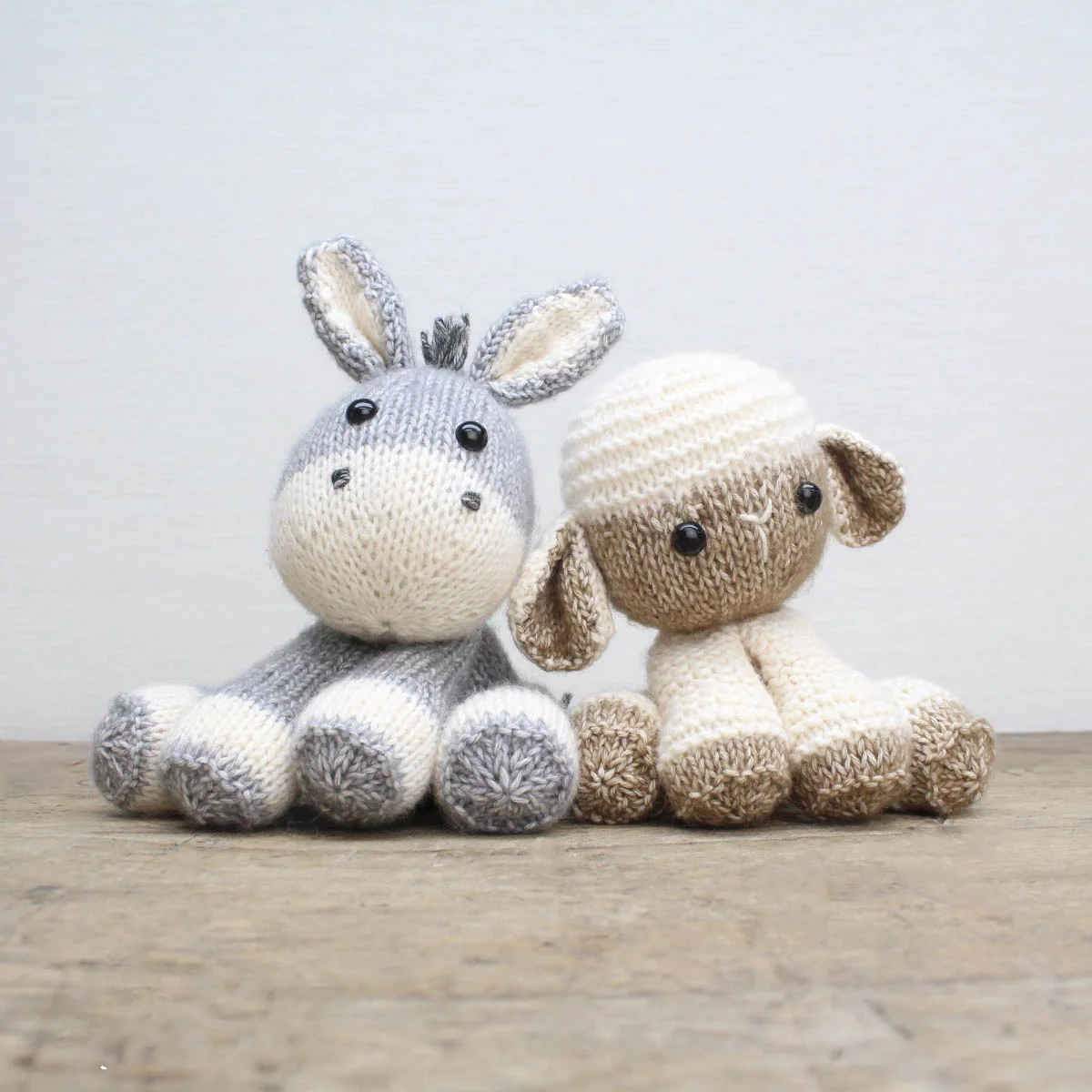 Knitted donkey and lamb soft toys sitting on a wooden surface with a light grey background