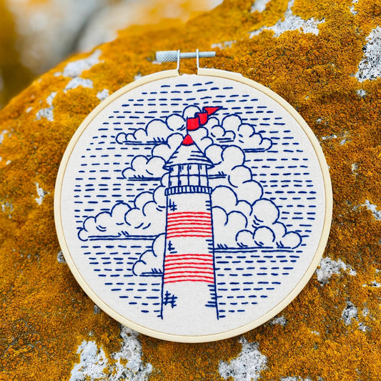 Finished embroidery of a red, white, and blue lighthouse on unbleached cotton, displayed in a wooden hoop on a lichen-covered stone