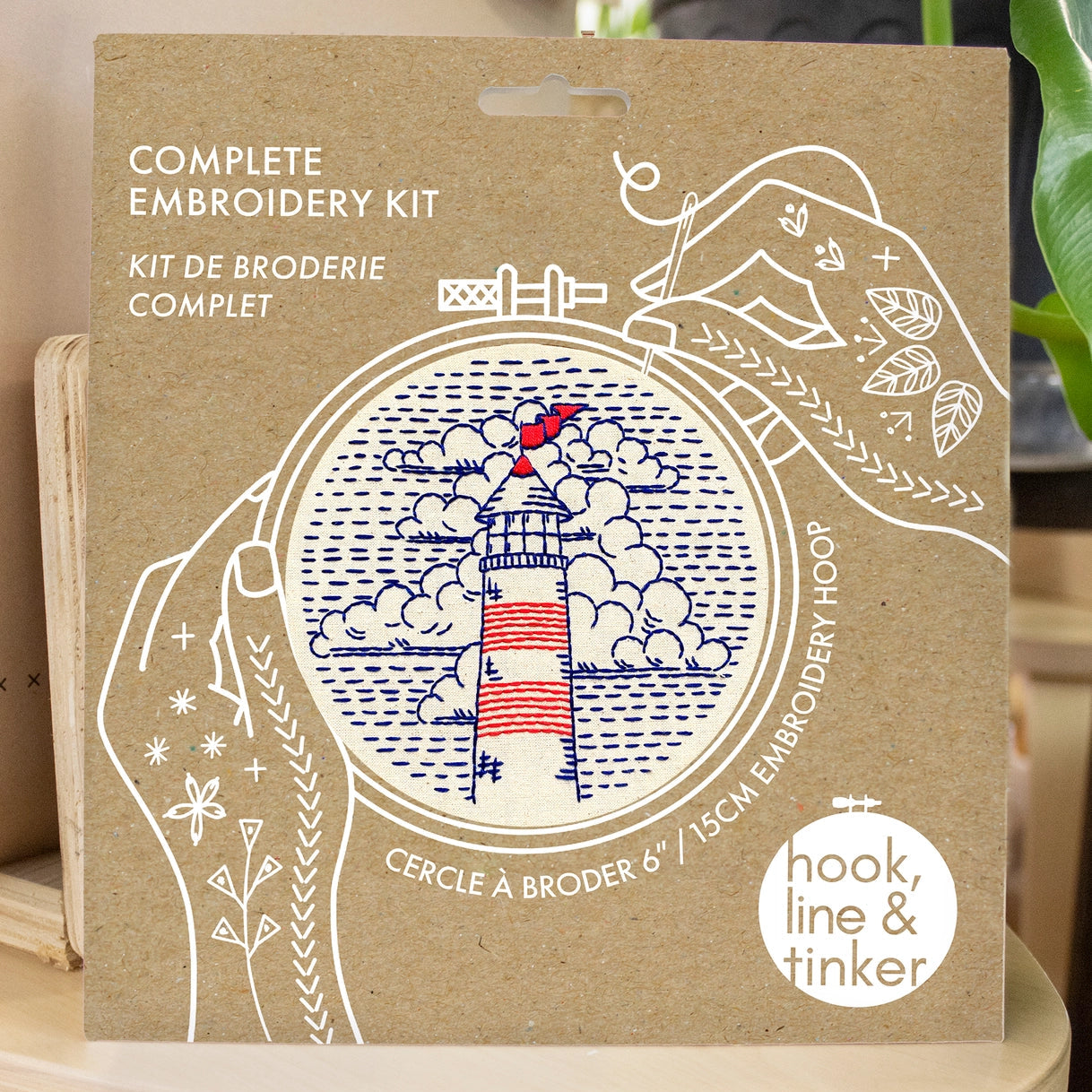 Kraft packaging for a lighthouse embroidery kit, showing illustrated hands holding a needle and thread around the design