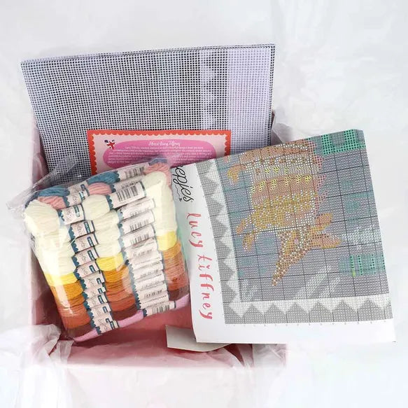 Cross-stitching materials including fabric, threads, and a pattern sheet in a box on a white background